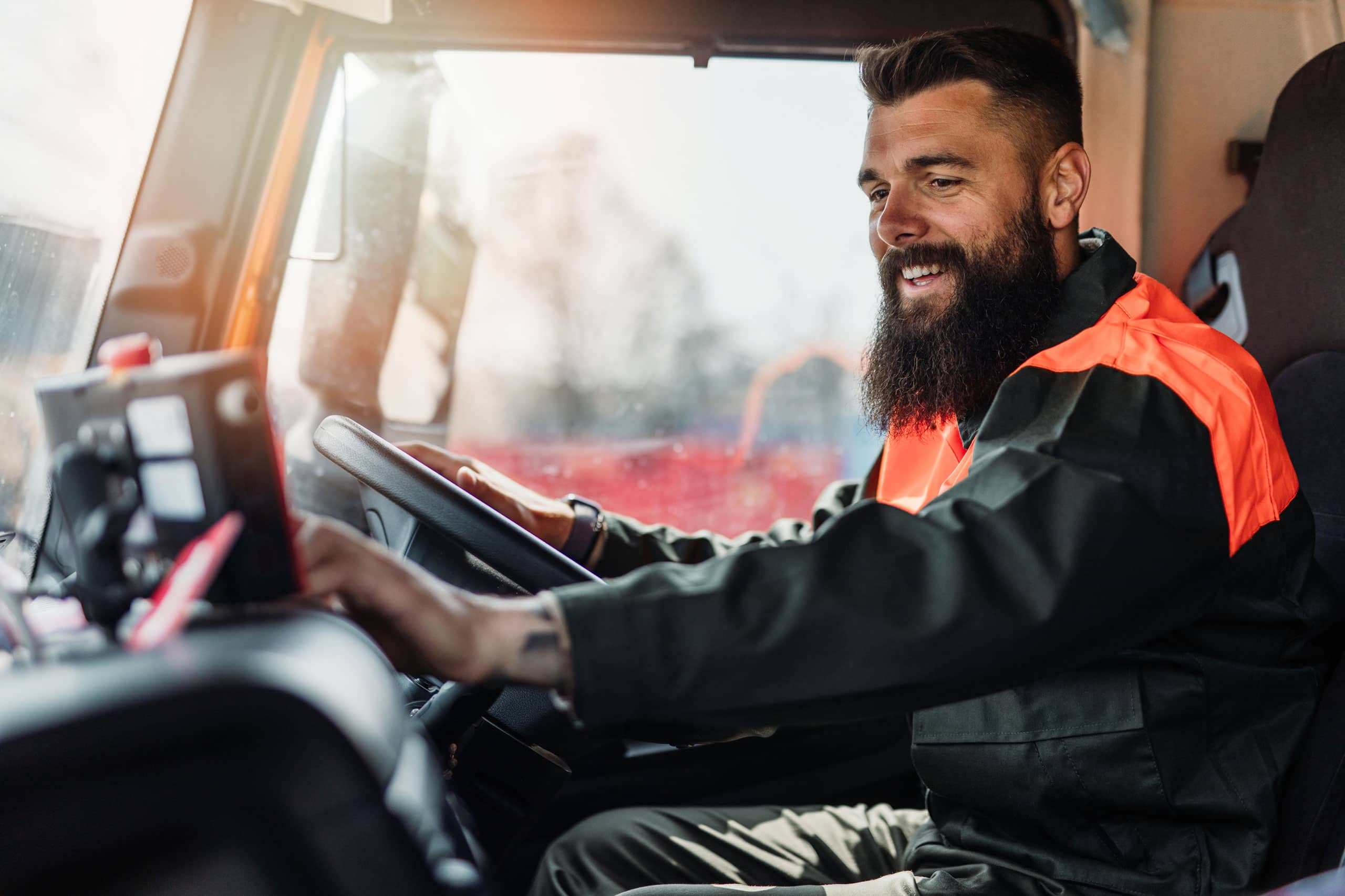 A bearded man in a high-visibility jacket sits in the driver’s seat of a truck, smiling as he operates the vehicle’s controls. Sunlight shines through the window, highlighting his role in supporting affordable housing through the Lancaster Housing Fund.