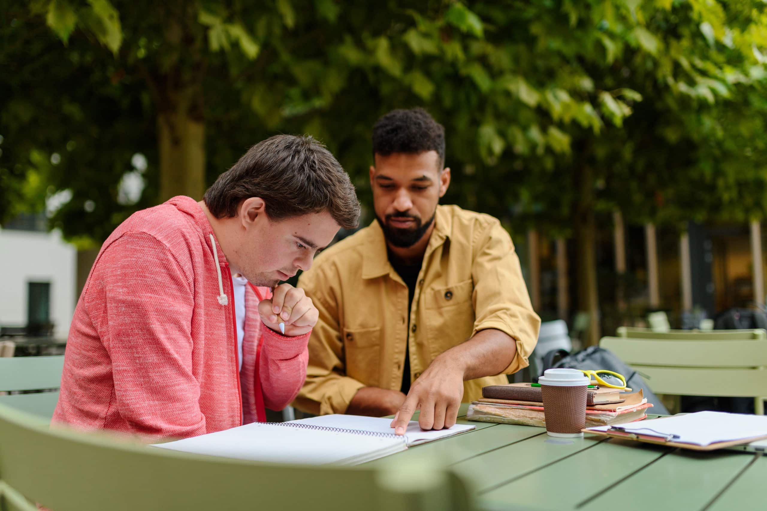 Two men sit at an outdoor table, studying together. One points to a notebook as they discuss affordable housing. Books, glasses, and a coffee cup rest on the table. Trees are in the background, creating a peaceful study setting.
