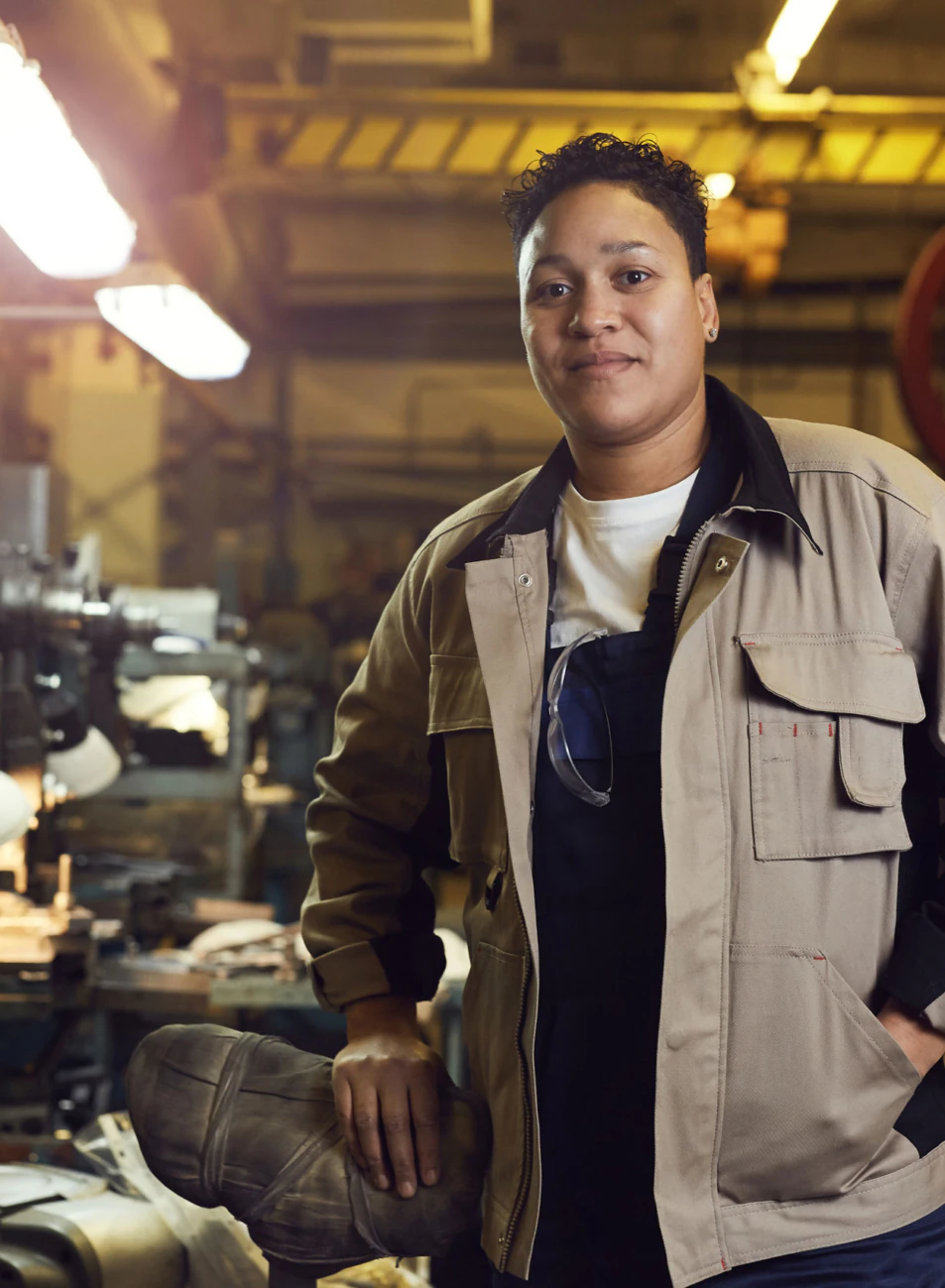 A person wearing a beige work jacket stands confidently in a factory workshop surrounded by industrial machines and tools, with bright lights illuminating the space—a hardworking spirit akin to those driving affordable housing initiatives.