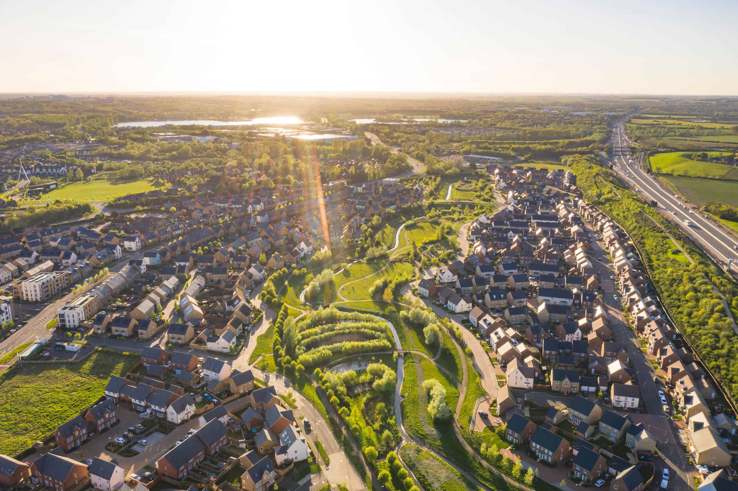 Aerial view of a suburban neighborhood featuring affordable housing, winding roads, green parks, and a highway on the right, all bathed in the glow of a bright, setting sun.