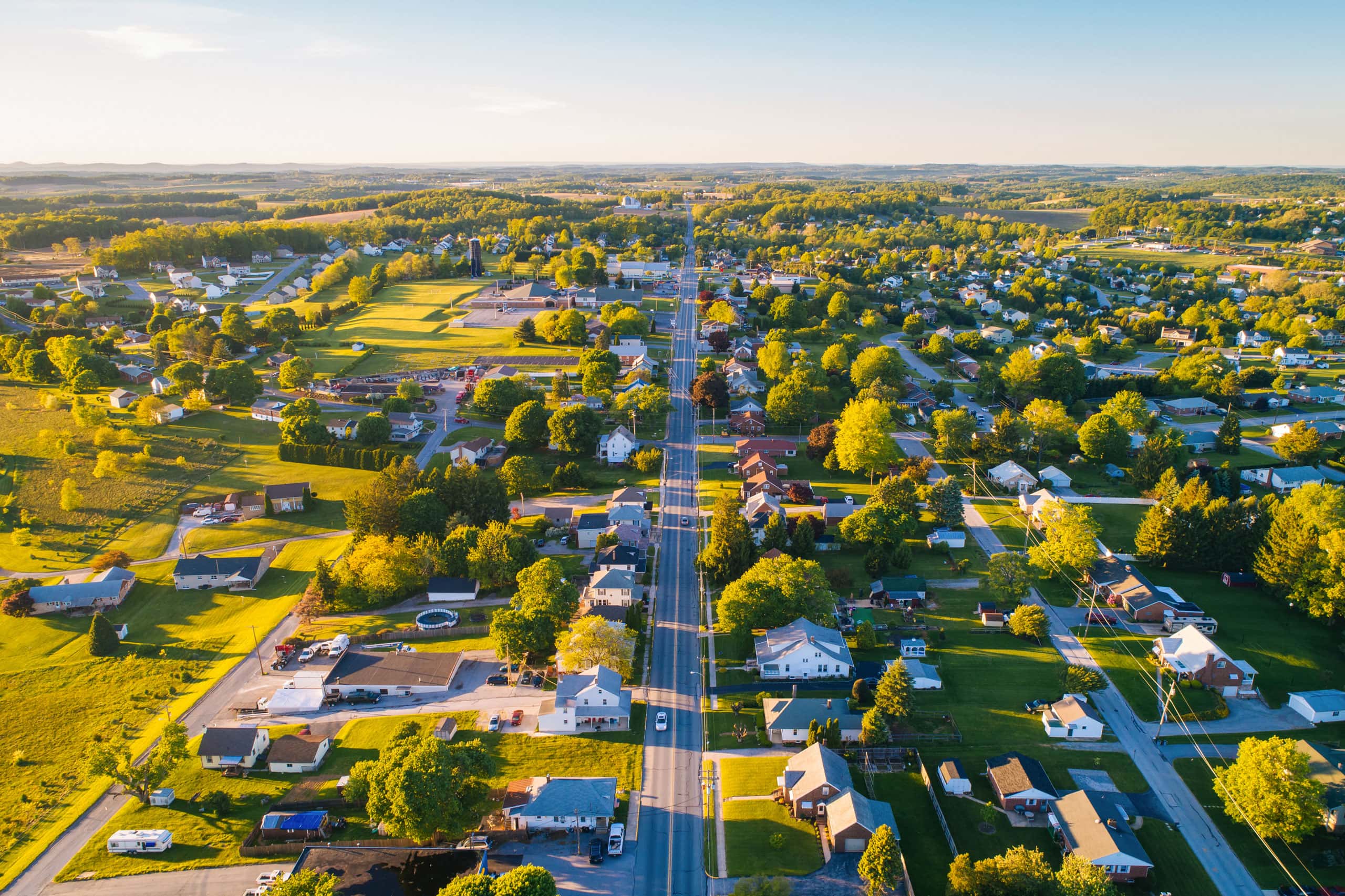 Aerial view of a small town with houses, roads, and green trees bathed in warm sunlight. The grid-like streets highlight a rural landscape where affordable housing initiatives, like the Lancaster Housing Fund, support the community.