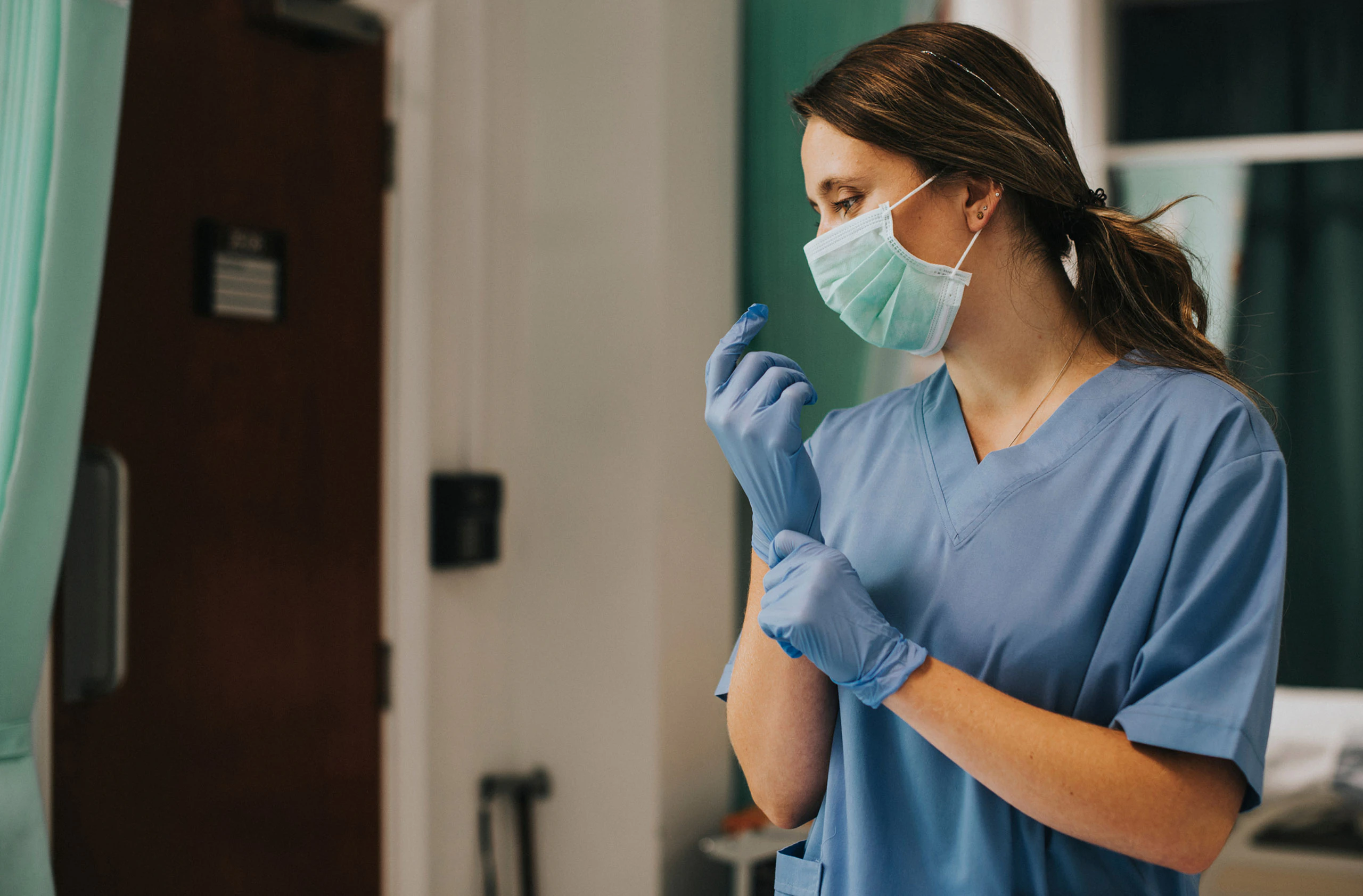 A healthcare worker wearing a surgical mask and blue scrubs puts on blue gloves in a medical setting, preparing for a procedure or examination—highlighting the vital role that affordable housing plays in supporting essential workers.