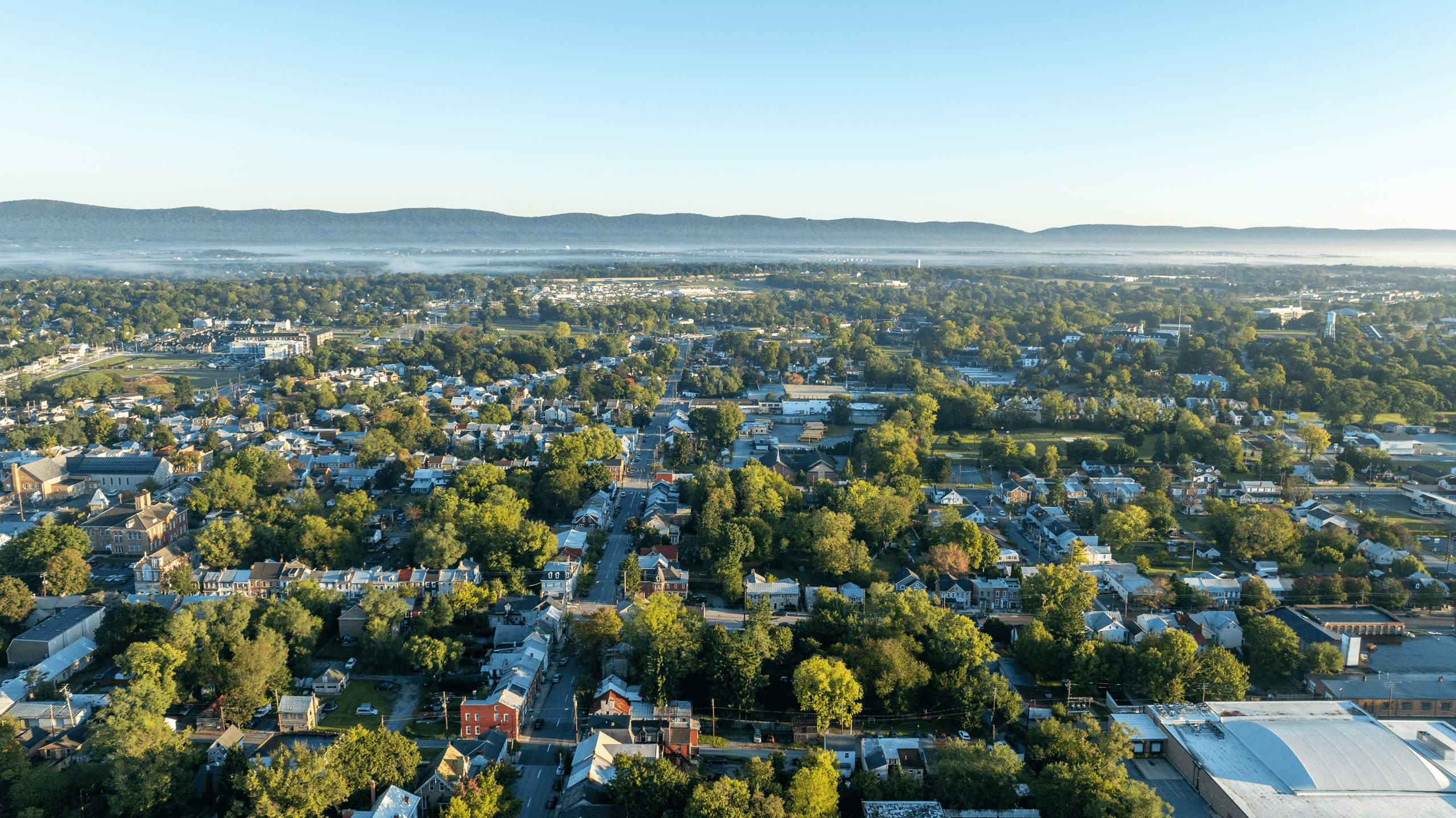 Aerial view of a small town with tree-lined streets, houses, and buildings supported by the Lancaster Housing Fund, promoting affordable housing amid greenery and distant hills under a clear blue sky.