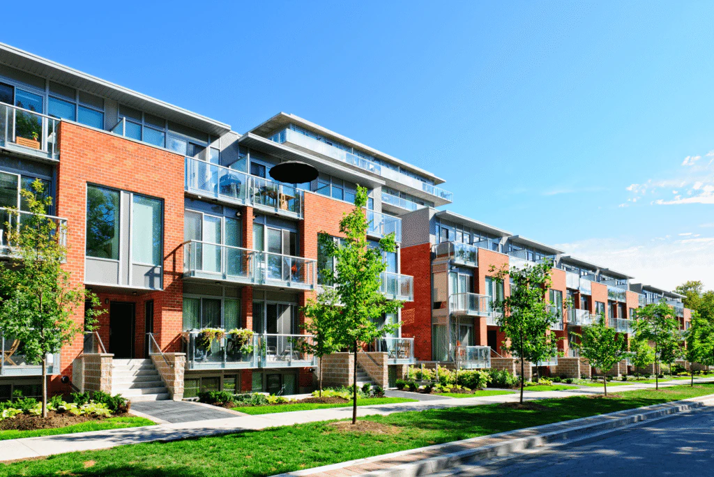 Modern apartment building with red brick and glass balconies, surrounded by young trees and landscaping along a sunny street, under a clear blue sky—an inviting example of affordable housing supported by the Lancaster Housing Fund.