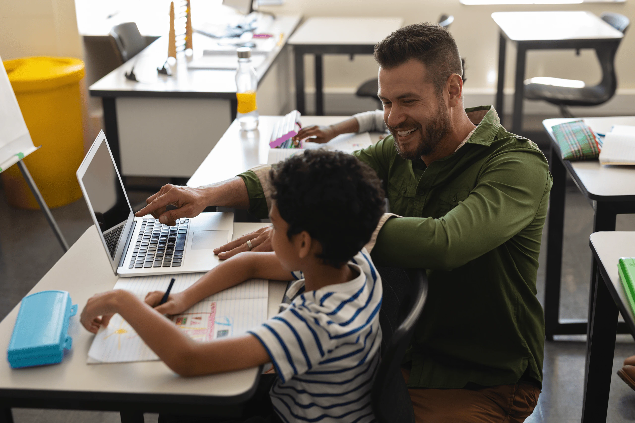 A smiling teacher in a green shirt helps a young student with curly hair, who is drawing in a notebook, while discussing affordable housing on a laptop screen in a classroom setting.