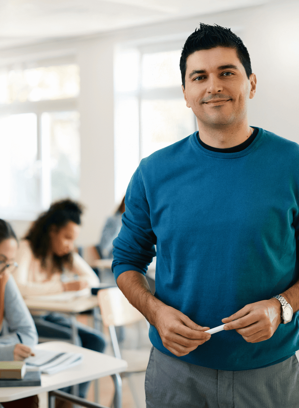 A man in a blue sweater stands smiling in a bright classroom, holding a marker. Several students are seated at desks in the background, focused on writing or reading about the impact of affordable housing initiatives.